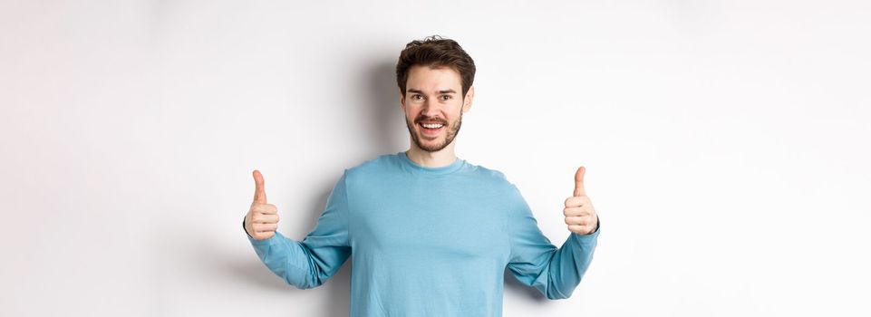 Cheerful young man showing thumbs up and smiling, recommend good product, praising choice, standing on white background.