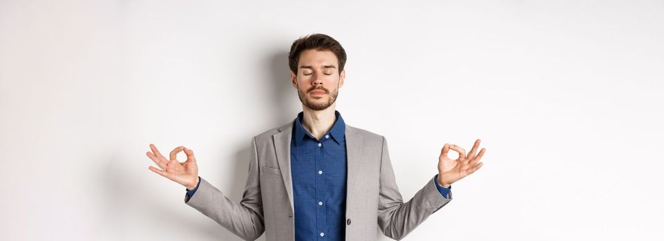 Calm and focused businessman meditating with eyes closed and hands spread sideways, finding peace in meditation, practice yoga breathing, standing on white background.