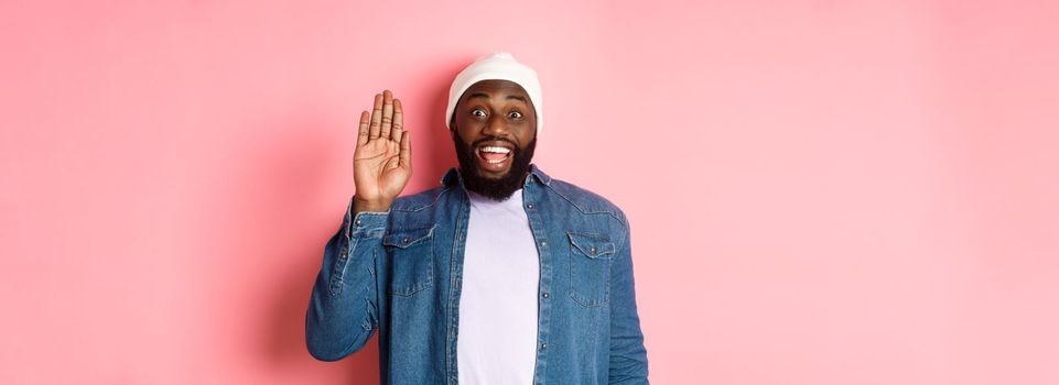 Friendly smiling Black man saying hello, waving hand, greeting you, standing over pink background.