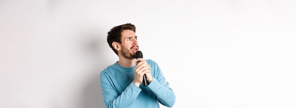 Worried and confused man reading lyrics on karaoke, looking left with unsure face, holding microphone and singing, white background.