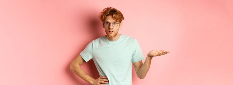 So what. Confused redhead man raising hand and shrugging, looking puzzled, dont understand something, standing over pink background.