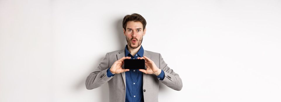 E-commerce and online shopping concept. Excited young man in business suit show empty smartphone screen and say wow amazed, white background.