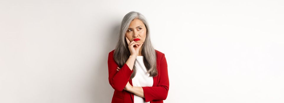 Troubled asian businesswoman in red blazer and lips, pouting and looking left annoyed, standing against white background.