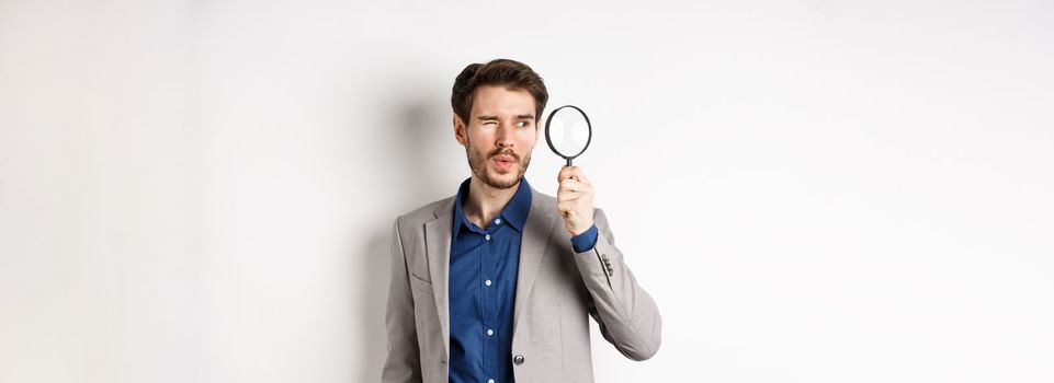 Handsome male model in suit looking through magnifying glass with interest, seeing something aside, white background.