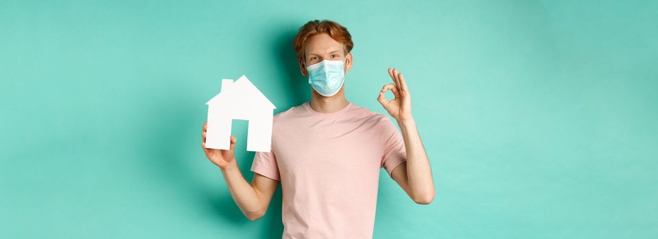 Covid-19 and real estate concept. Cheerful guy in face mask showing house cutout and okay sign, standing over turquoise background.