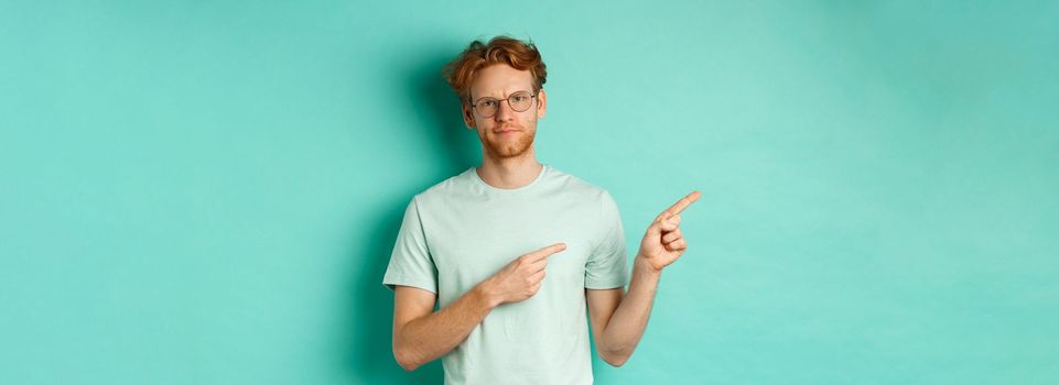 Judgemental redhead man in glasses grimacing, frowning disappointed and pointing left, showing bad promo offer, standing over turquoise background.