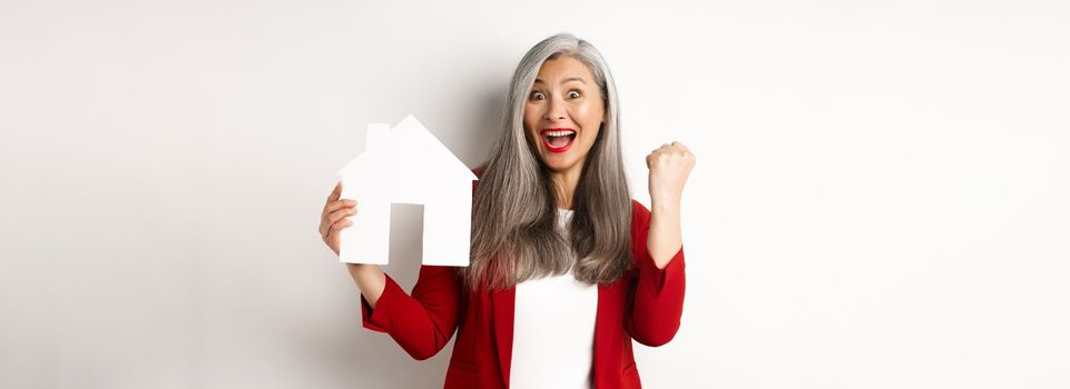 Happy asian grandmother showing paper house cutout and fist pump gesture, scream yes with joy, buying property, standing over white background.