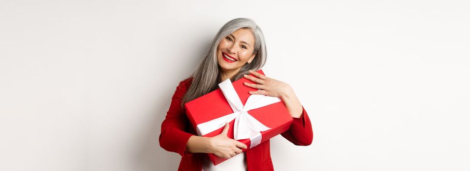 Happy asian grandmother hugging red gift box and smiling grateful, thanking for present, standing over white background.