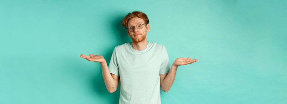 Portrait of confused redhead man in t-shirt and glasses know nothing, shrugging shoulders and looking clueless at camera, standing against turquoise background.