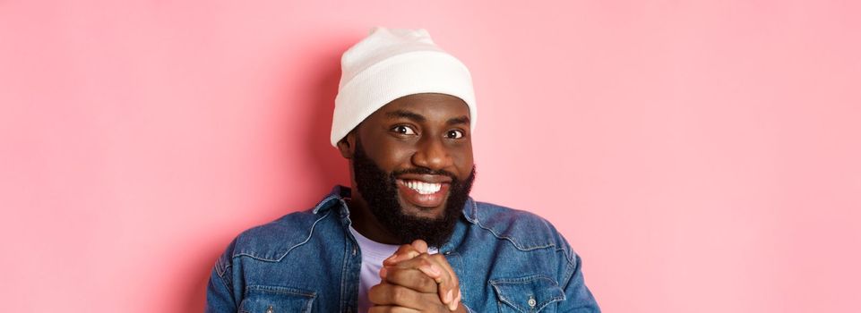 Close-up of devious and happy african-american man smiling satisfied, clasp hands think something good, expect profit, standing over pink background.