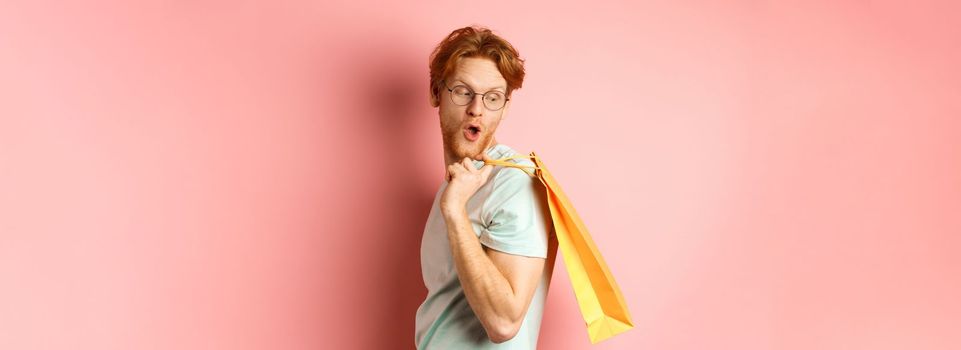 Carefree young man with red hair and glasses, walking with shopping bag, looking behind his shoulder with amazed expression, standing over pink background.