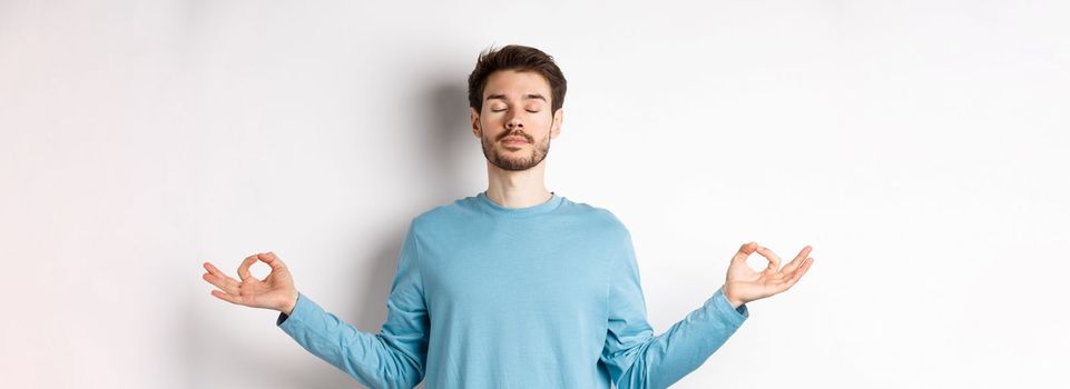 Young man trying to relax in meditation, standing calm with stretch out hands in zen mudra gesture, practice yoga on white background.