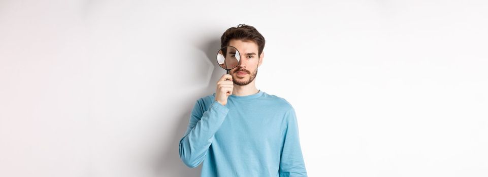 Man searching for something, looking through magnifying glass at camera, standing on white background.