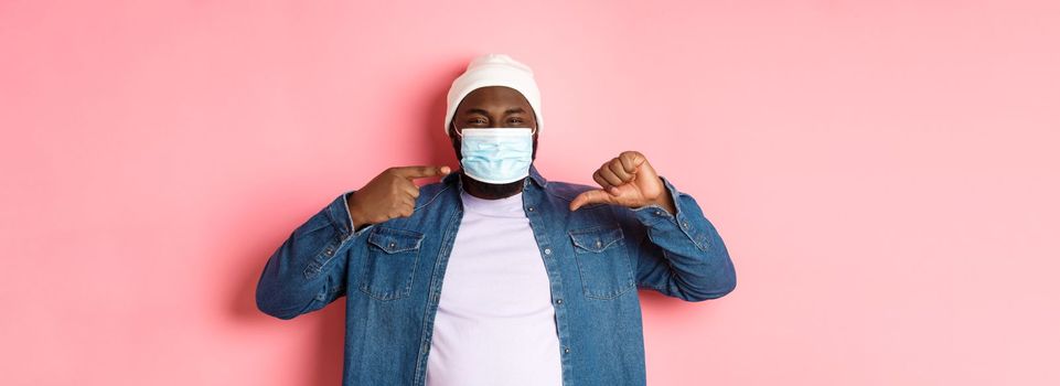 Coronavirus, lifestyle and global pandemic concept. Displeased hipster guy pointing at face mask, showing thumbs-down, dislike wearing it, pink background.