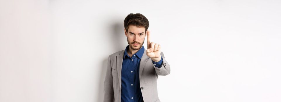 Listen to me. Serious male ceo manager showing one finger, scolding or telling to stop, tell no, hold right there sign, standing on white background in suit.