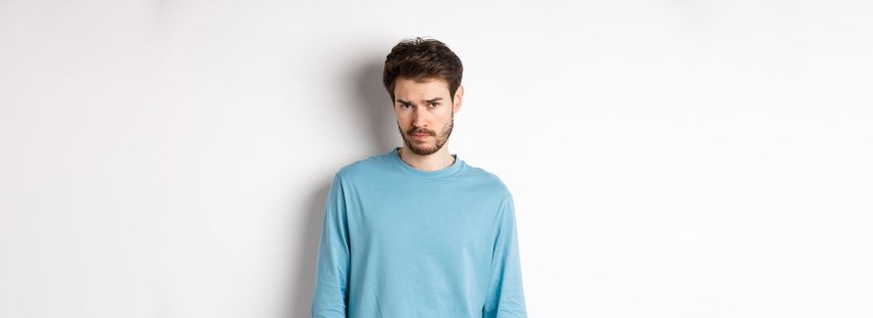 Sad young bearded man sulking, looking upset at camera, offended by someone, standing against white background.