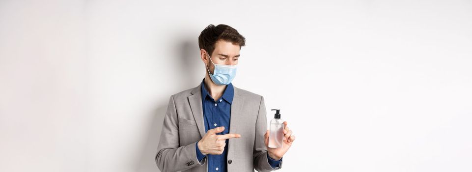 Covid-19, pandemic and business concept. Office worker in medical mask pointing at bottle of hand sanitizer, using antiseptic at work, white background.
