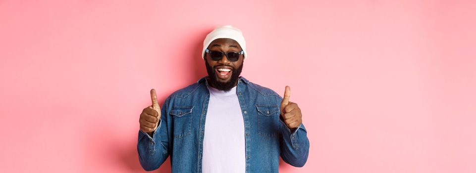 Satisfied african-american hipster guy in beanie and sunglasses showing thumbs-up, approve and praise good choice, smiling pleased, pink background.