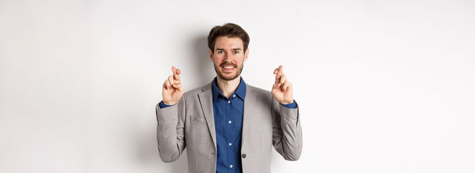 Hopeful smiling businessman looking optimistic, cross fingers good luck, praying to win, waiting for results with hope, standing on white background.