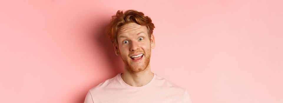 Headshot of funny redhead guy showing tongue, making silly faces at camera, standing joyful against pink background. Copy space