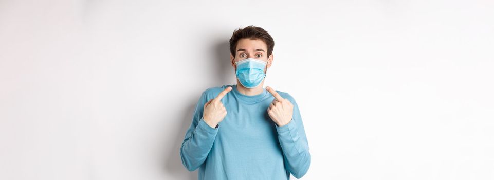 Coronavirus, health and quarantine concept. Confused guy pointing at his medical mask and looking surprised at camera, standing over white background.