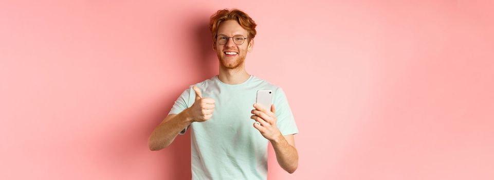 Happy young man with red messy hair, showing thumbs-up while using mobile phone, smiling and praising application, standing over pink background.