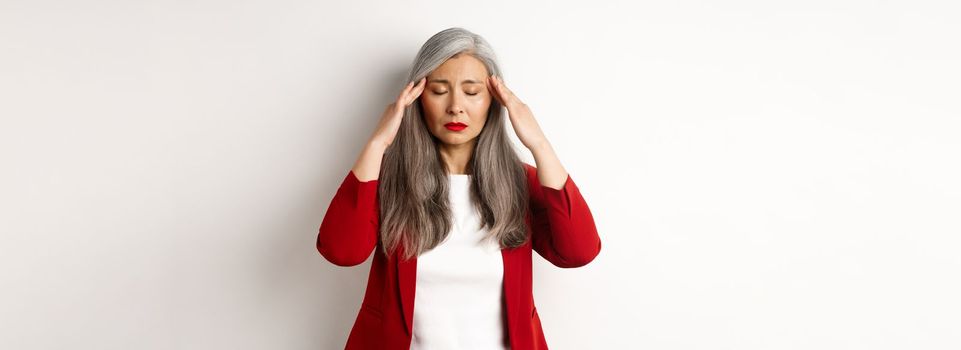 Distressed asian businesswoman trying to calm down, massaging temples on head to soothe headache, standing over white background.