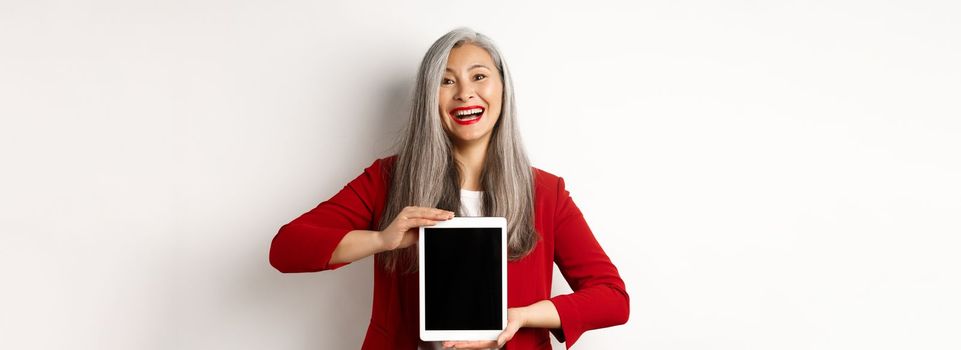 Business. Cheerful asian businesswoman in red blazer, showing digital tablet screen and smiling, introduce app or promotion, white background.