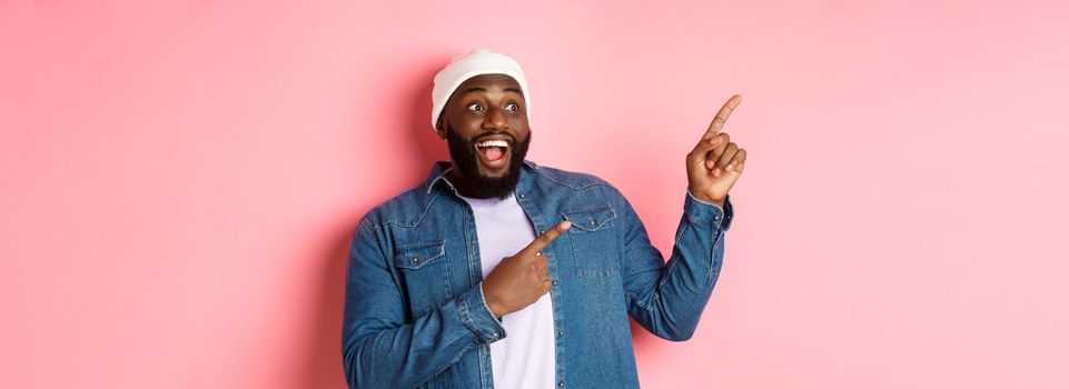 Amazed Black guy showing awesome promo offer, smiling and pointing fingers at upper left corner banner, pink background.