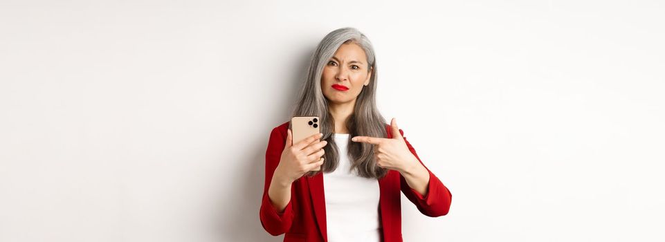 Disappointed mature asian woman complaining, pointing finger at smartphone and looking displeased, standing over white background.
