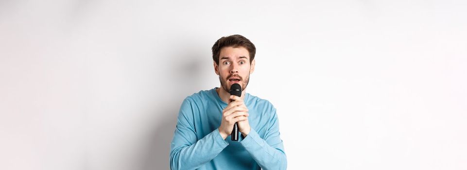 Confused young man looking nervously at camera while singing karaoke, holding microphone, standing over white background.