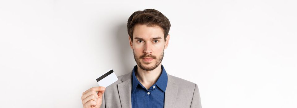 Finance. Close up of handsome bearded businessman in suit showing plastic credit card, standing serious on white background.