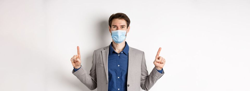 Covid-19, pandemic and business concept. Happy businessman in medical mask and suit pointing fingers up, standing on white background.