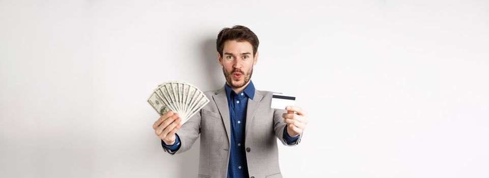 Happy successful businessman in suit showing money and credit card, smiling at camera, white background.