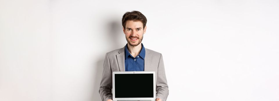 Handsome caucasian businessman in suit showing empty laptop screen, demonstrate promo, standing on white background.