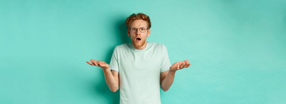 Confused and shocked guy with red messy hair and glasses, shrugging and raising hands, staring at something strange, cant understand, standing over turquoise background.
