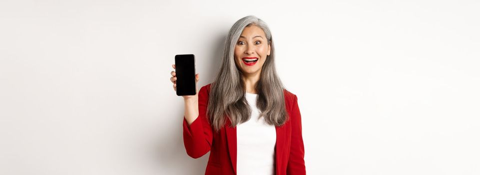 Cheerful asian female entrepreneur showing mobile phone screen, smiling amused, standing over white background.