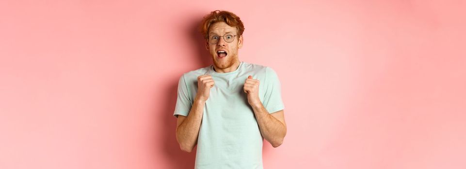 Portrait of scared redhead guy in glasses staring startled at camera, press hands to body and scream of fear, standing over pink background.