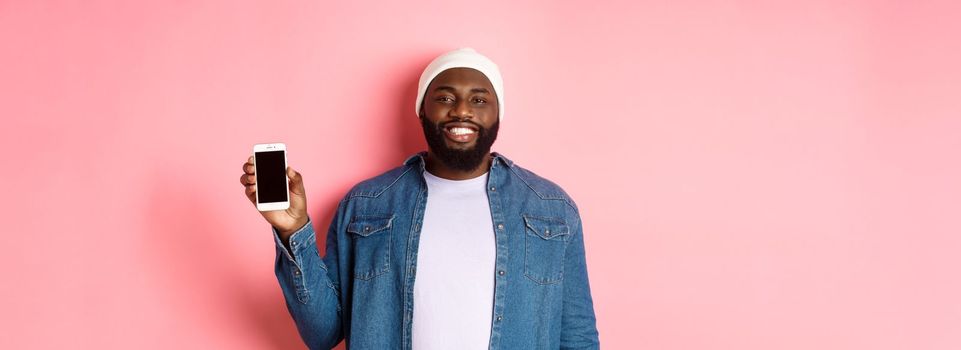 Handsome hipster guy in beanie and denim shirt smiling, showing mobile phone screen with happy face, introduce application, standing over pink background.