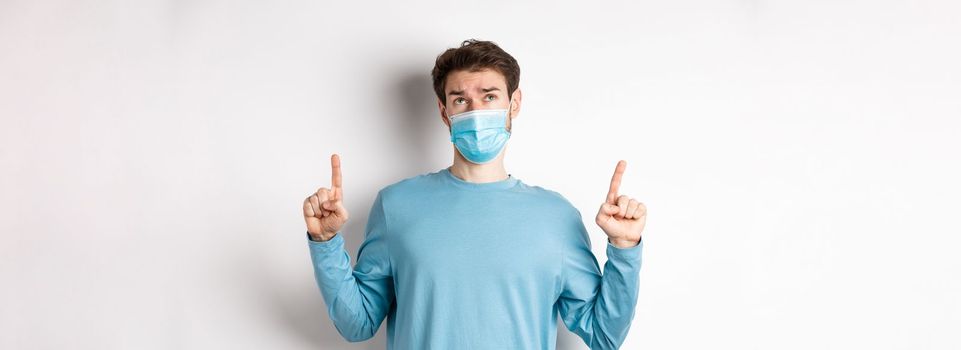 Coronavirus, health and quarantine concept. Skeptical and doubtful man in medical mask pointing, looking up, feeling indecisive, standing over white background.