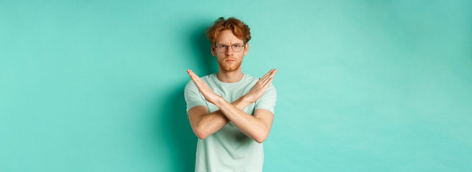 Serious and confident redhead man in t-shirt and glasses saying no, showing cross gesture to stop you, refucing or declining something, standing over turquoise background.
