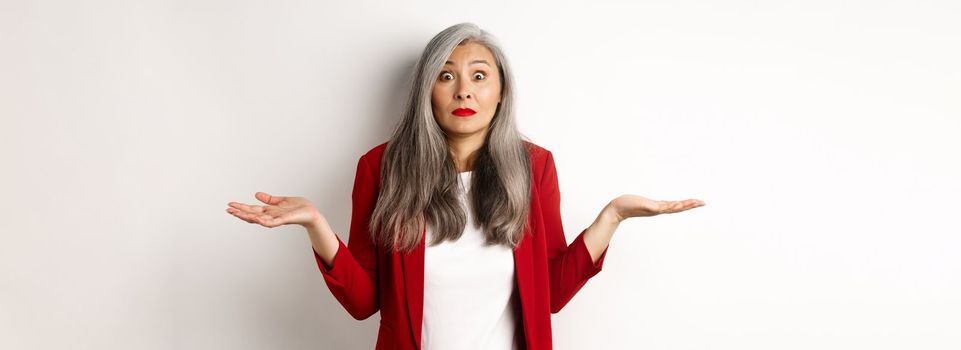 Confused asian senior woman shrugging, spread hands sideways and staring questioned at camera, dont know anything, standing over white background.