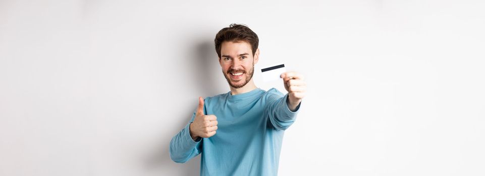 Handsome caucasian man showing plastic credit card with thumb-up, recommend and praise good bank offer, standing on white background