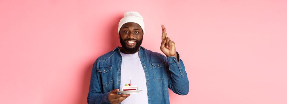 Handsome african-american guy celebrating birthday, making wish with fingers crossed, holding bday cake with candle, standing against pink background.