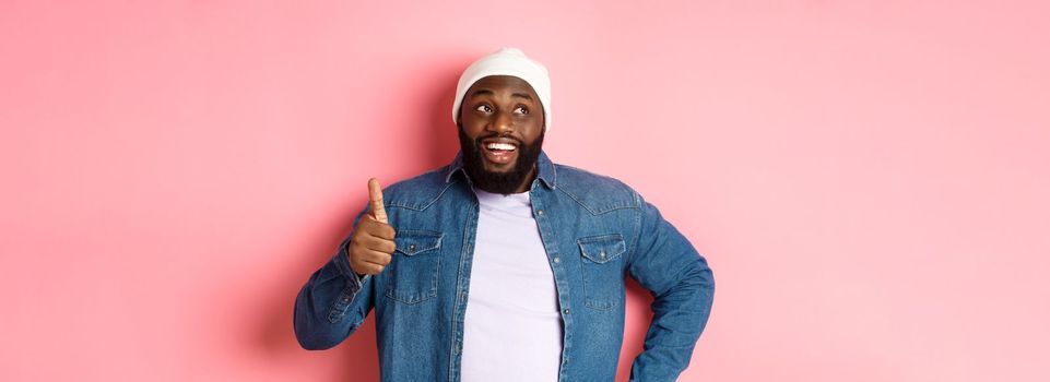 Happy Black man showing thumb-up, looking at upper left corner thoughtful, standing over pink background.