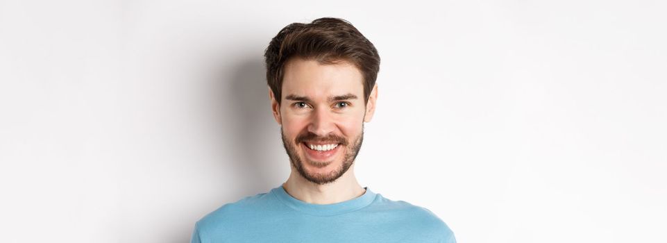 Close-up of handsome caucasian man smiling with white teeth, looking confident at camera, standing in blue shirt on white background.
