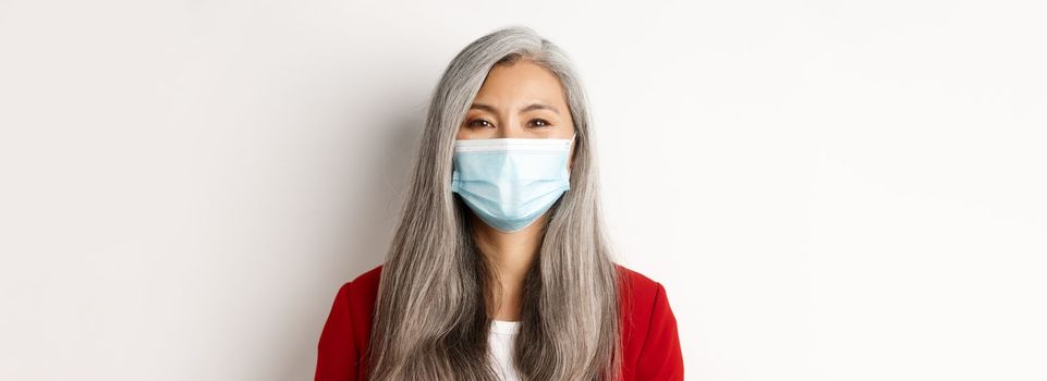 Covid, pandemic and business concept. Close-up of cheerful asian senior businesswoman in medical mask smiling at camera, white background.