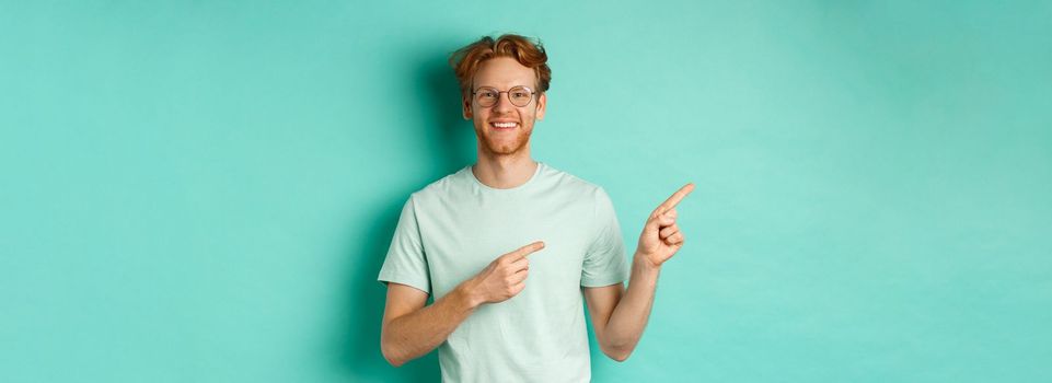 Young cheerful man with short red hair and glasses, pointing fingers left at copy space, smiling white teeth, showing advertisement, mint background.