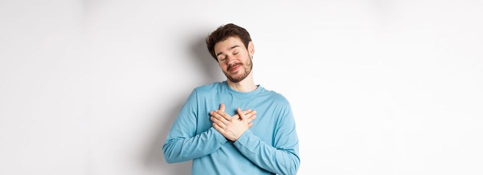 Handsome young man feeling romantic, holding hands on heart and thinking about good pleasant memories, standing over white background.