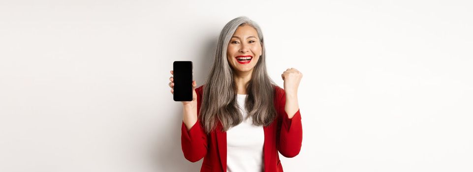 Success. Asian senior woman showing smartphone blank screen and fist pump, winning prize online, standing over white background.